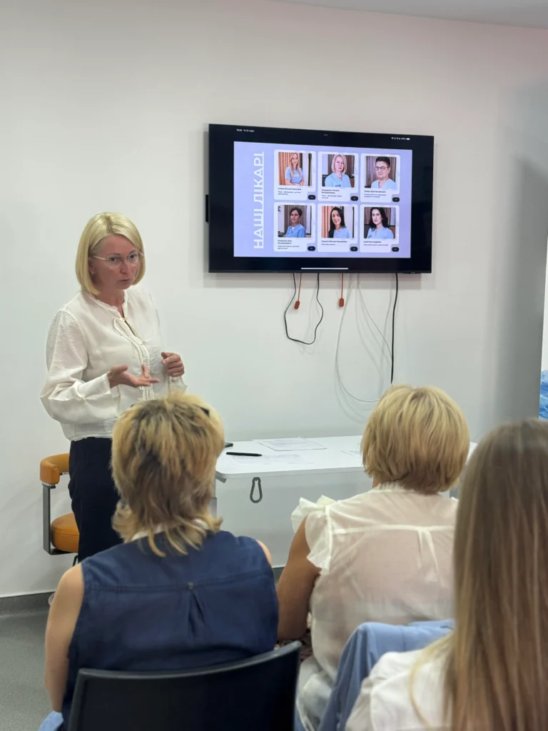 Лікарка виступає перед учасниками конференції; на екрані показані фотографії команди лікарів.